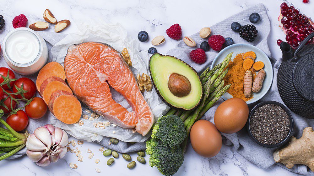 Spread of food: tomatoes, sweet potato, fish, avocado, broccoli, eggs, asparagus, ginger, chia seeds, edamame, raspberries, blackberries, blueberries, pomegranate, almonds, walnuts, and oats
