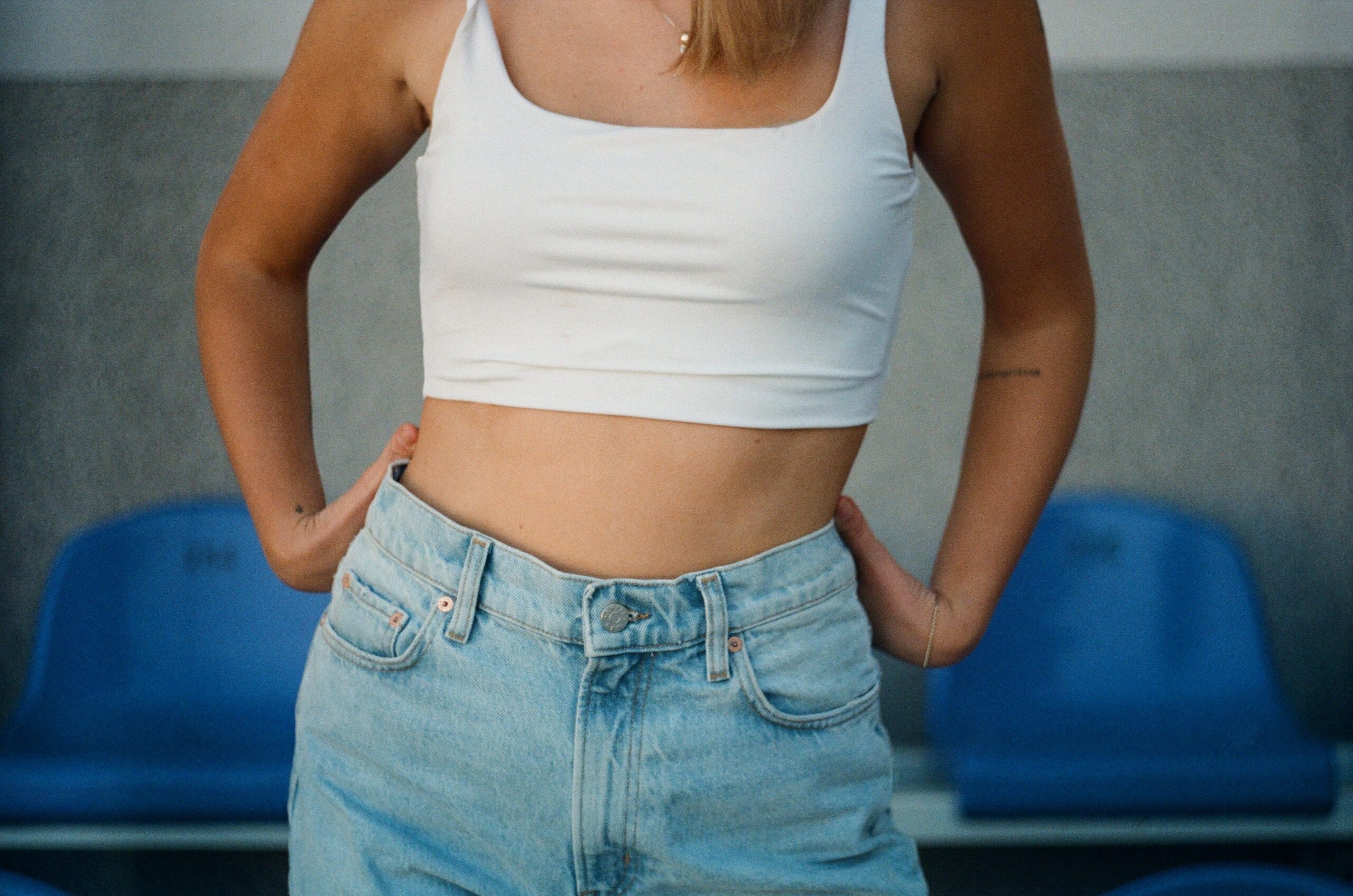 Person wearing white cropped tank top with light blue denim shorts. Their hands are resting on their hips.